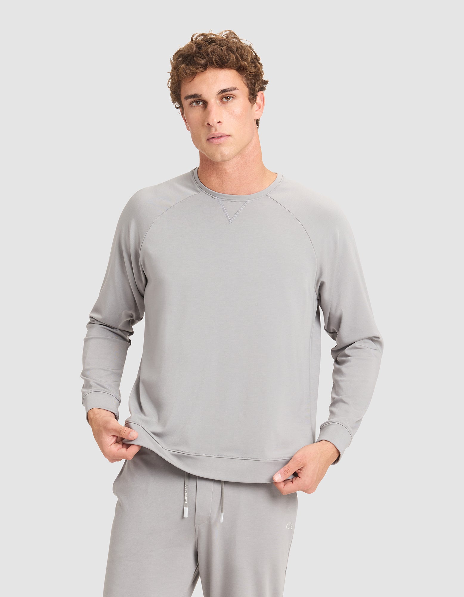 A young man with curly hair wears Cozy Earth's Men's Brushed Bamboo Pullover Crew in light gray, standing against a plain gray background and holding the hem of his sweatshirt. 