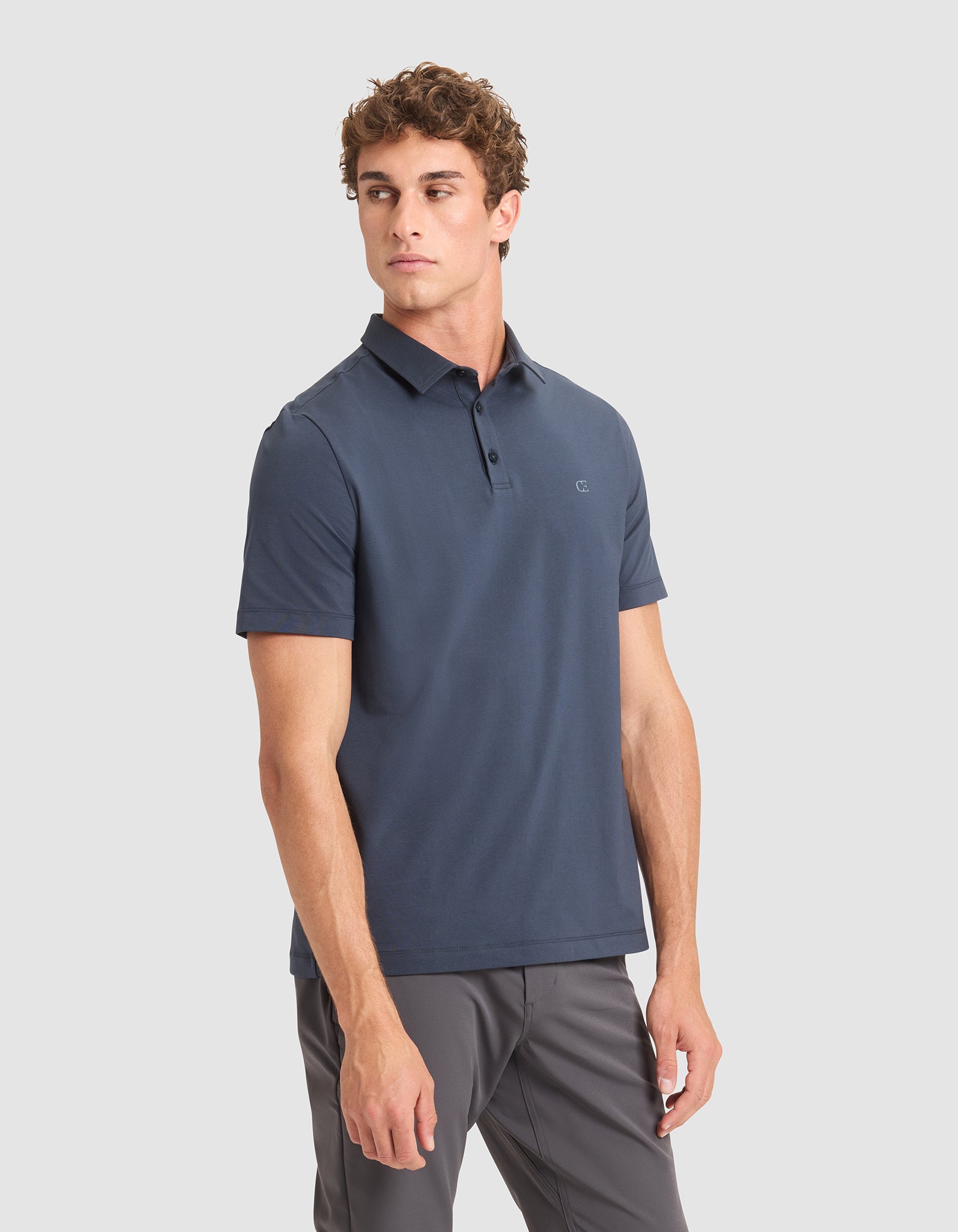 A man with short curly hair wears the Cozy Earth Men's Everyday Polo in dark blue and gray pants, standing against a plain light gray background and looking slightly left with a neutral expression. 