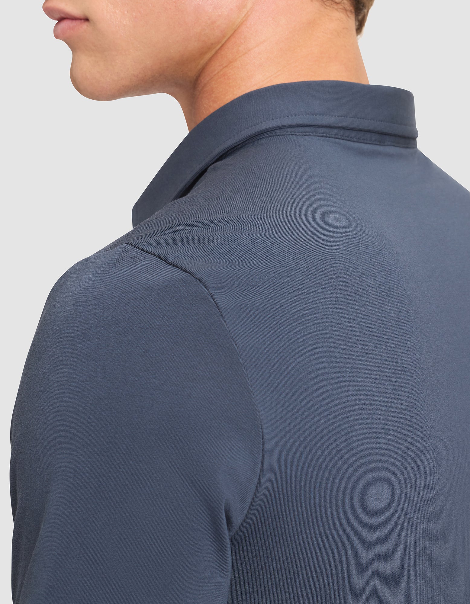 A close-up of a person wearing the Cozy Earth Men's Everyday Polo in dark blue, shown from the side and back. The image highlights the fabric, collar, and shoulder details with part of the head visible. 
