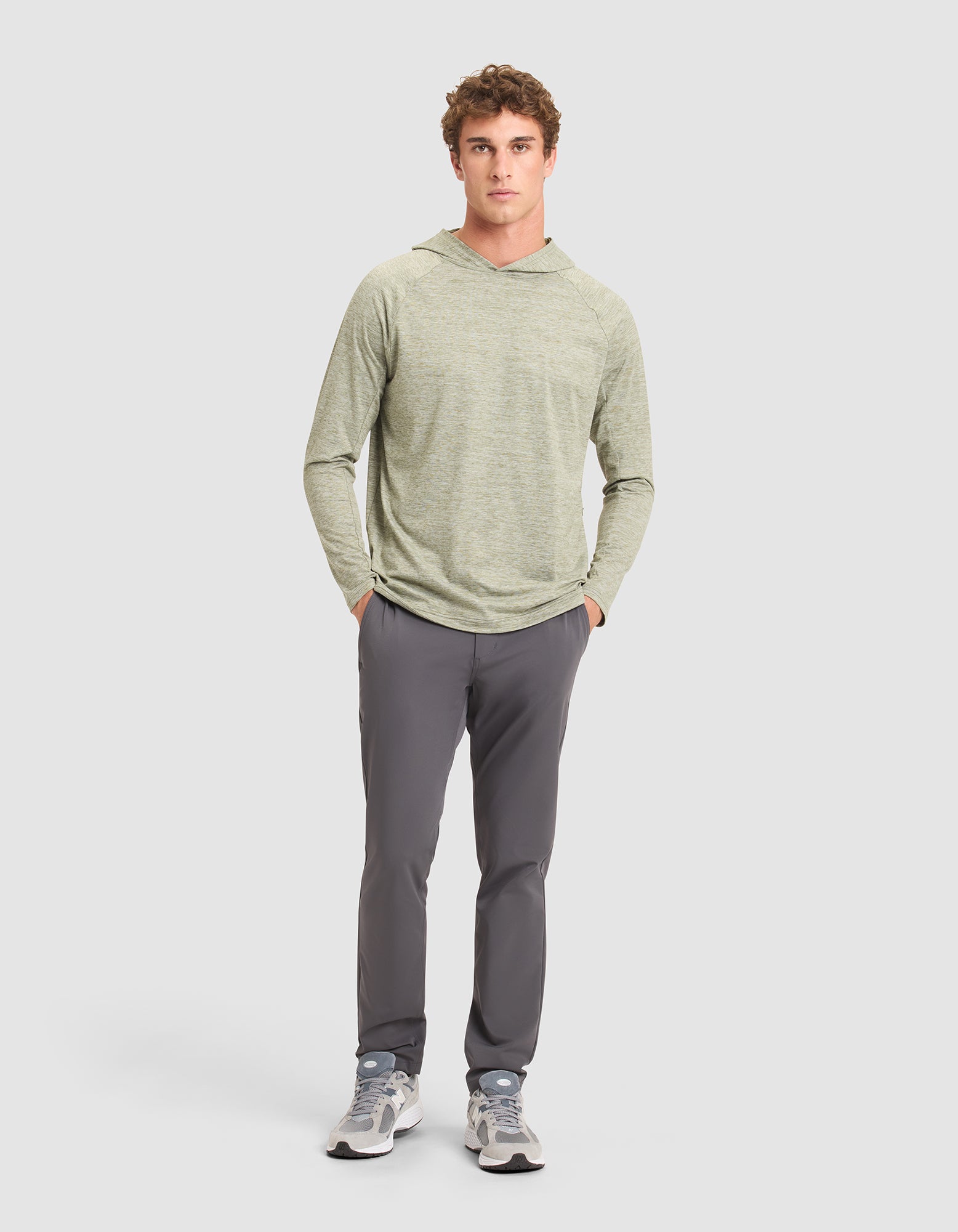 A young man stands against a plain background wearing the Cozy Earth Men's Lightweight Performance Hoodie in light green, paired with gray pants and gray sneakers, with his hands in his pockets. 