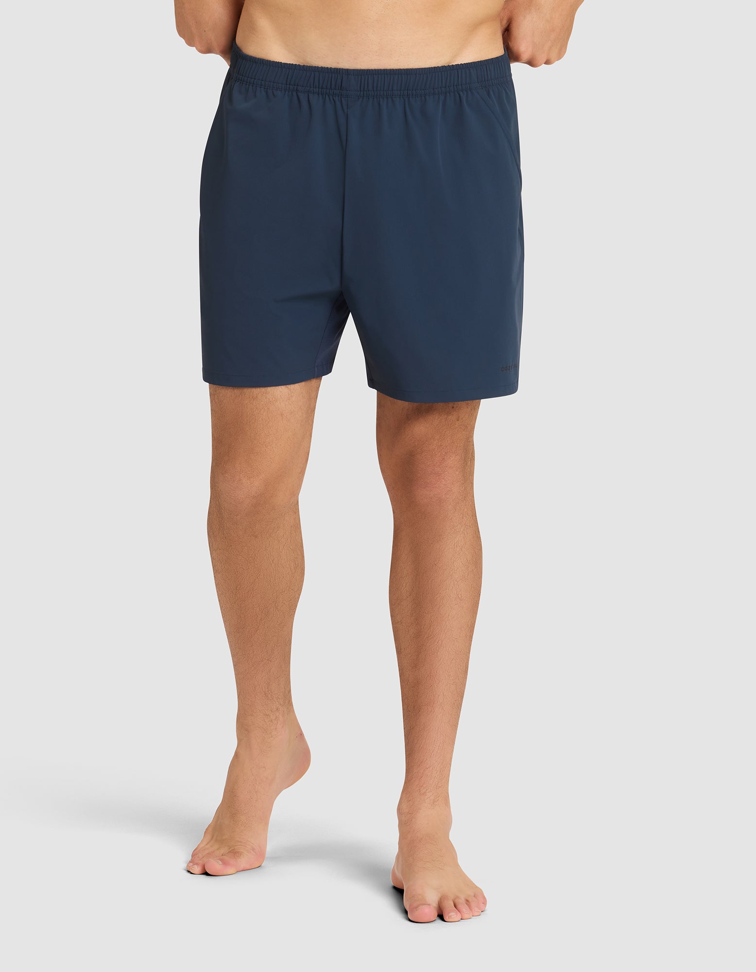 A person stands barefoot against a plain light background, wearing Cozy Earth's Men's Performance Sleep Short in dark blue with an elastic waistband. The upper body and head are not visible. 