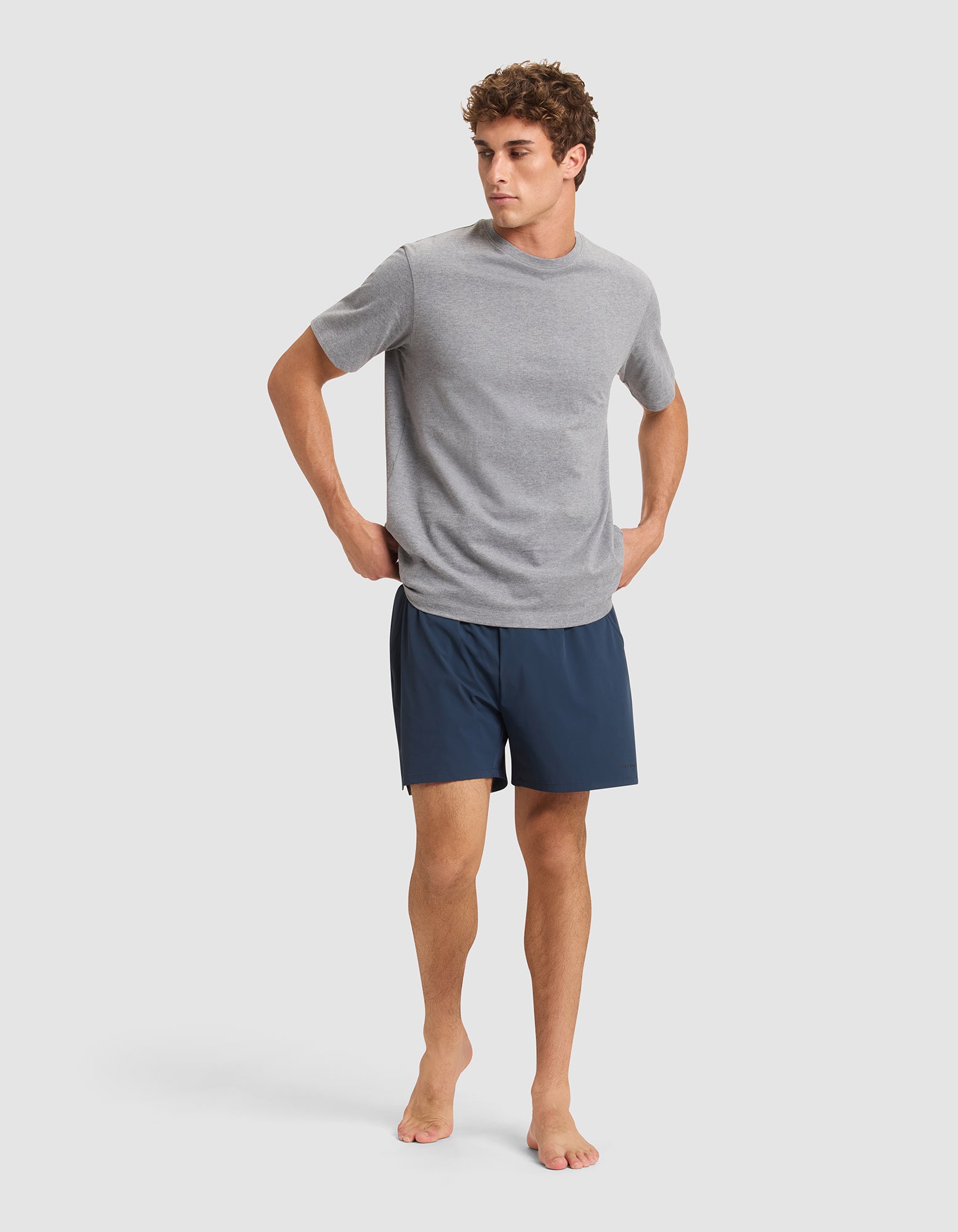 A man stands barefoot against a plain background, wearing a light gray short-sleeve T-shirt and Cozy Earth Men's Performance Sleep Short in dark blue, with his hands resting on his hips. 