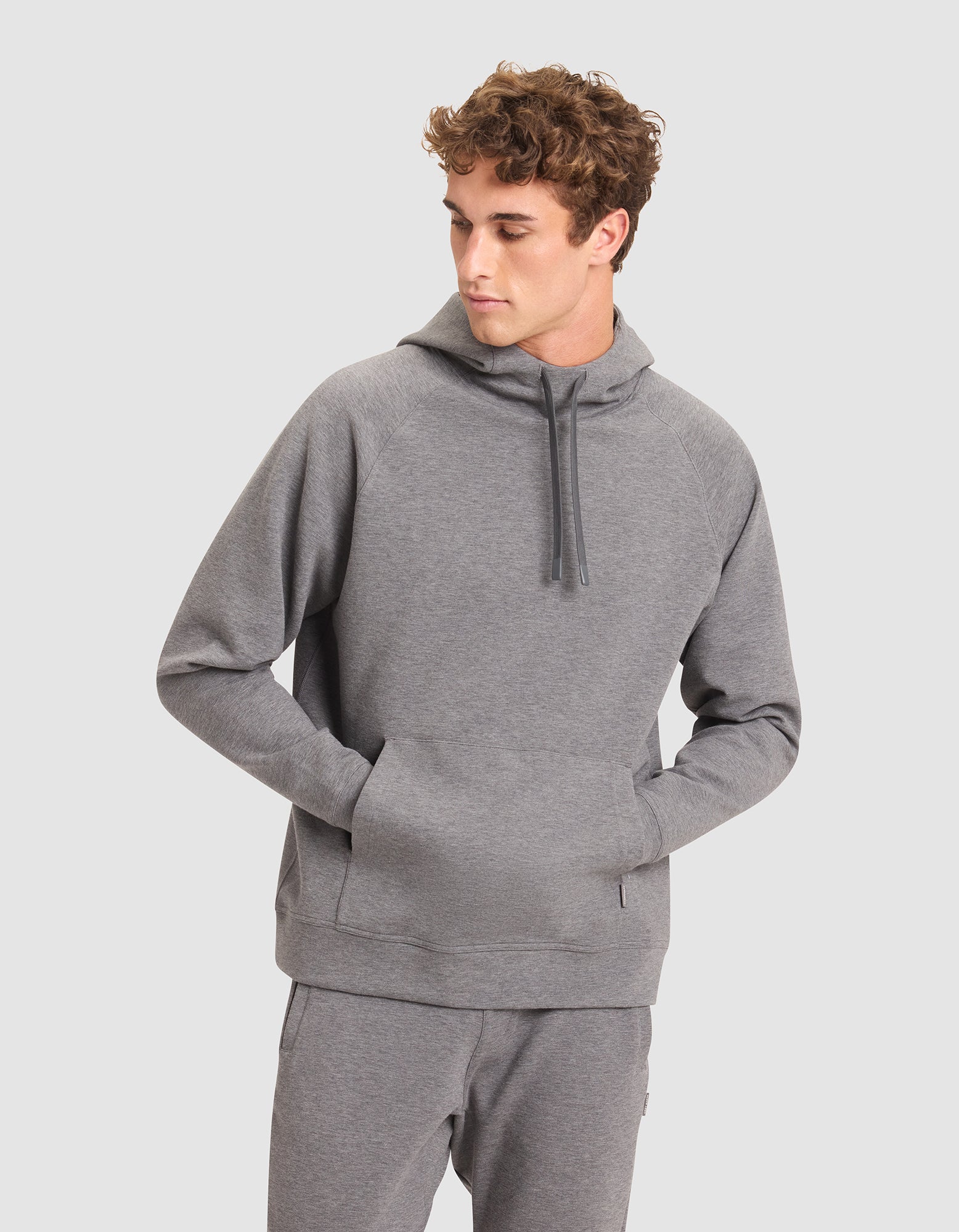 A young man wears the Cozy Earth Men's StretchTech Hoodie in light gray with matching sweatpants, hands in the hoodie pocket, looking left against a plain light gray background. 