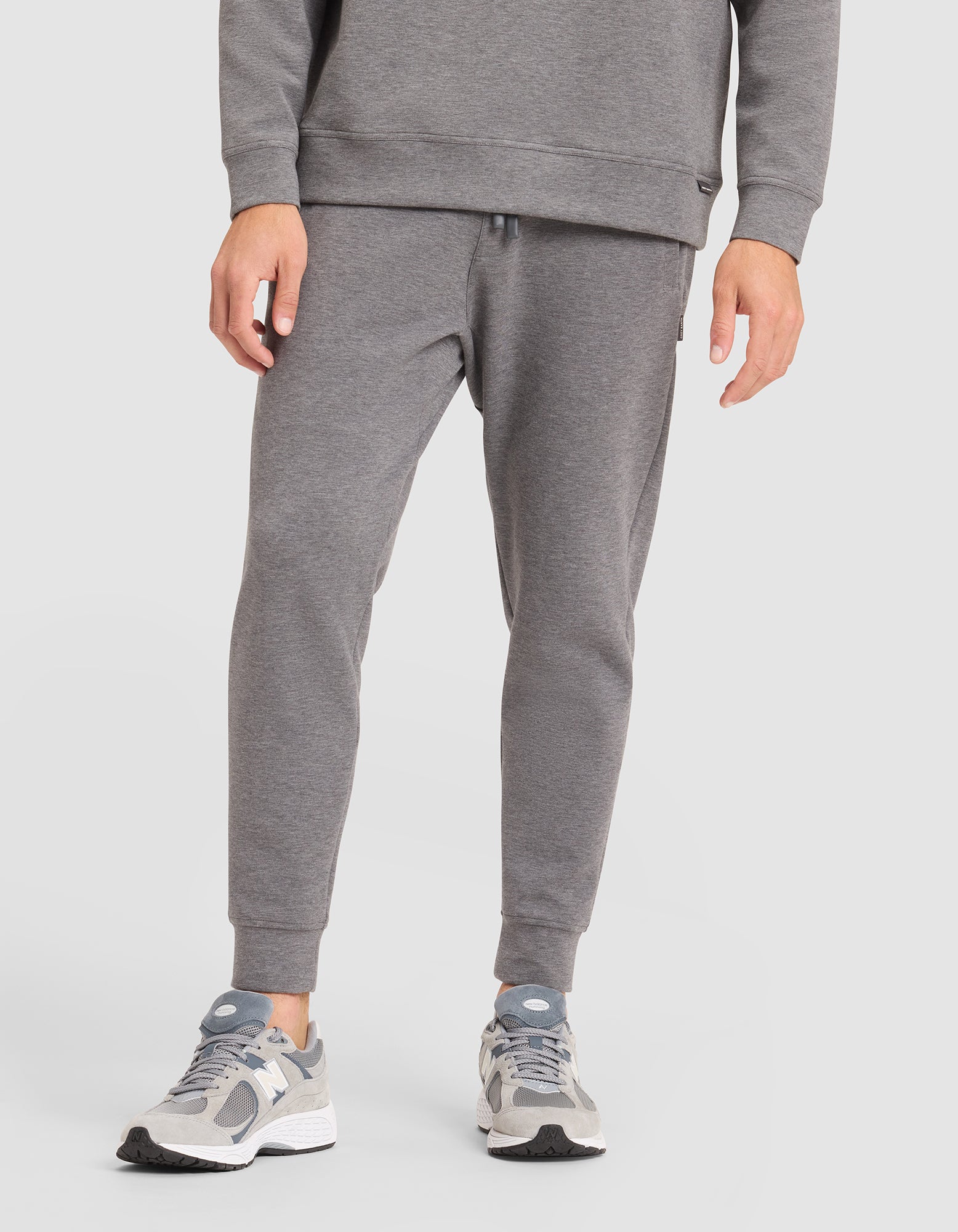 A person is shown from the waist down, wearing Cozy Earth's Men's StretchTech Jogger in gray with a matching sweatshirt and gray-white sneakers, standing against a plain white background. 