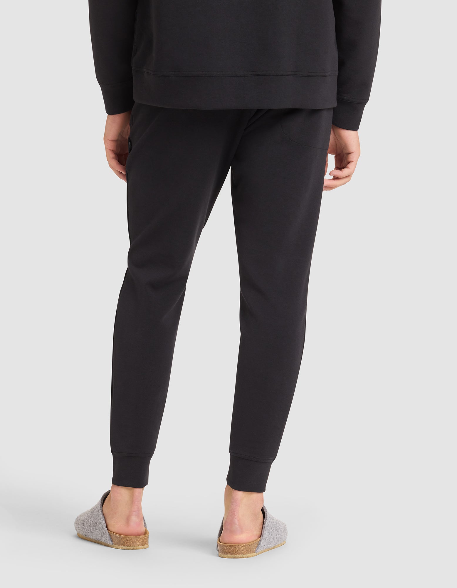 A person stands with their back to the camera, wearing Cozy Earth Men's StretchTech Jogger pants and a matching black sweatshirt, paired with light gray slippers with cork soles. The background is plain light gray. 