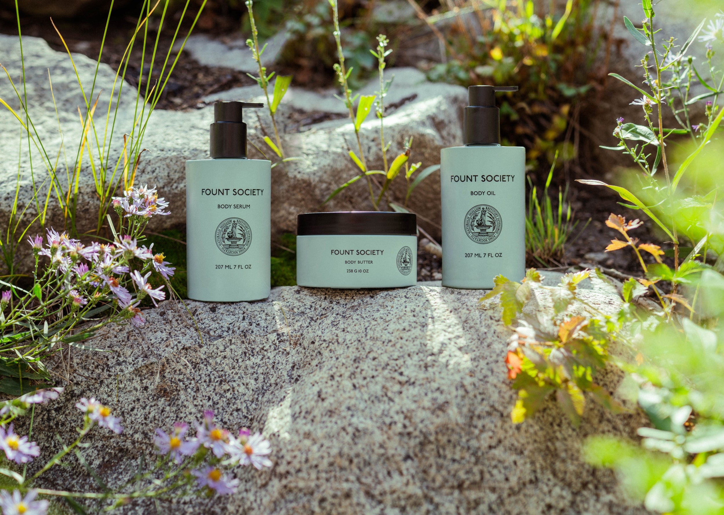 Three Cozy Earth skincare products from the Moisture Trio, including body serum, body polish, and body oil, are displayed outdoors on a rock amidst greenery and blooming wildflowers. The natural setting highlights the organic essence of the products.
