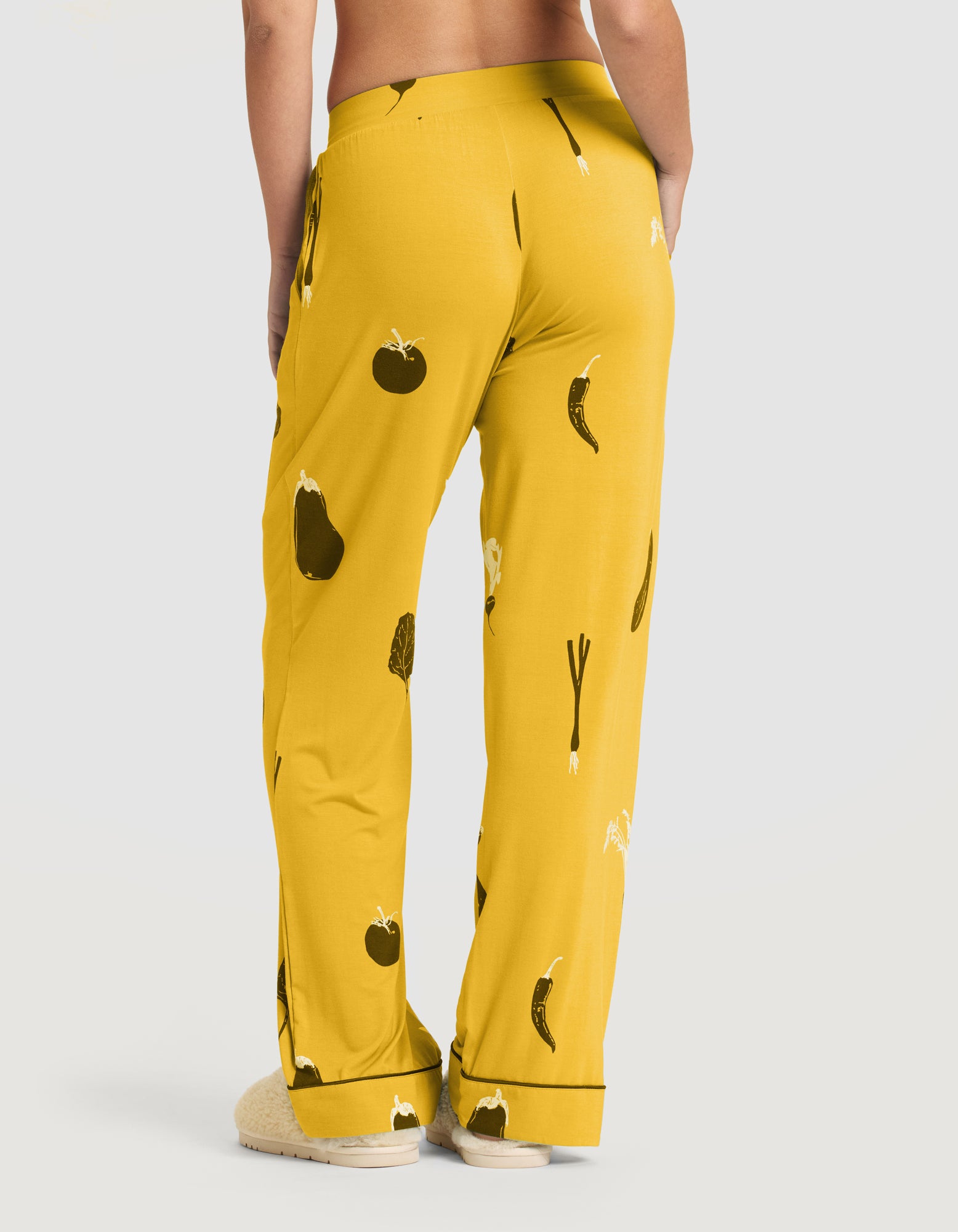 A person, barefoot, stands with their back to the camera in a plain light gray setting, showcasing the Cozy Earth Women's Bamboo Stretch-Knit Short Sleeve & Pant Pajama Set (Veggie), featuring yellow pants with black and white vegetable prints. 