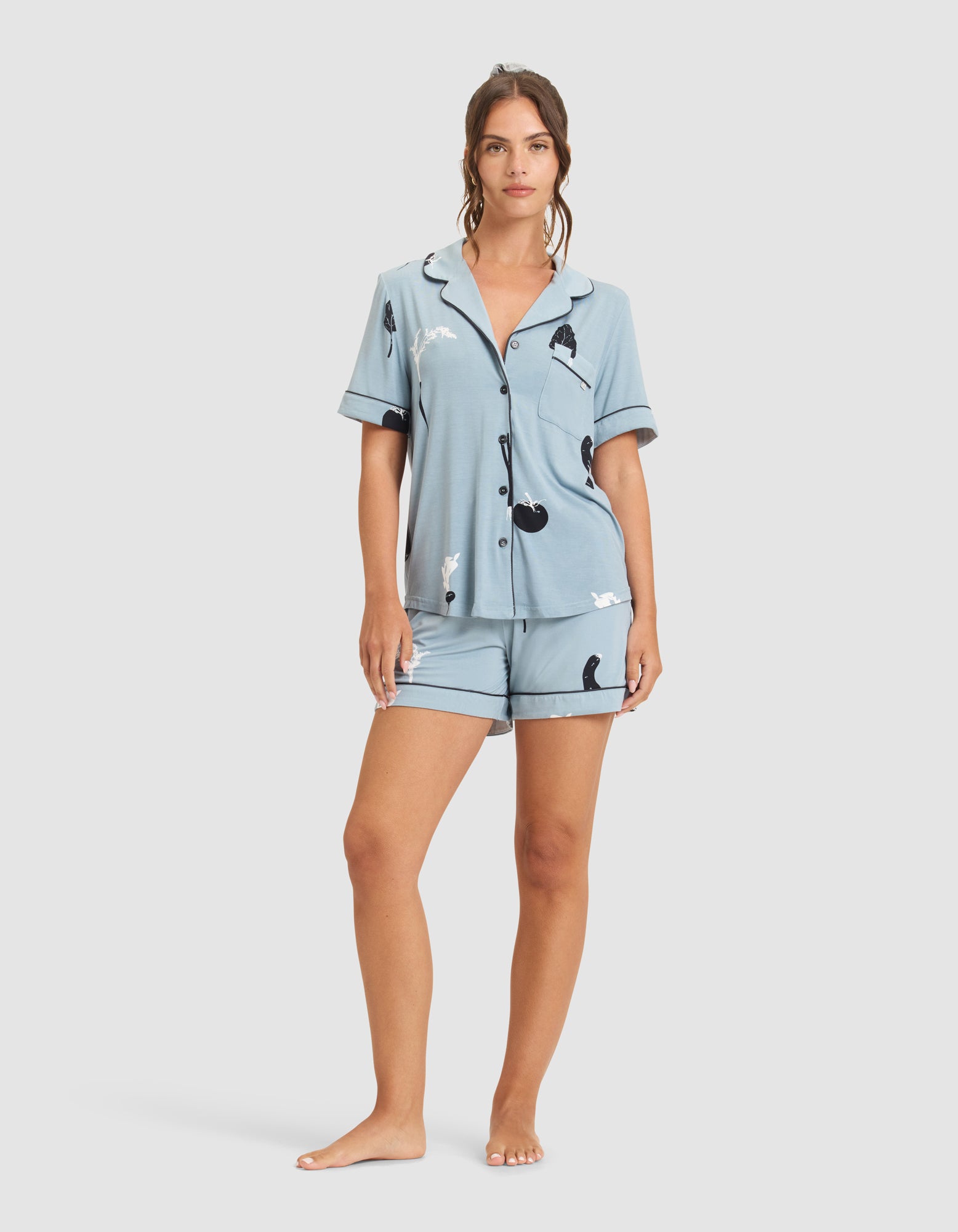 A woman stands barefoot facing forward against a white background, wearing the HIDE Women's Bamboo Stretch-Knit Short Sleeve Pajama Top and matching shorts, both featuring a playful black and white animal print. 