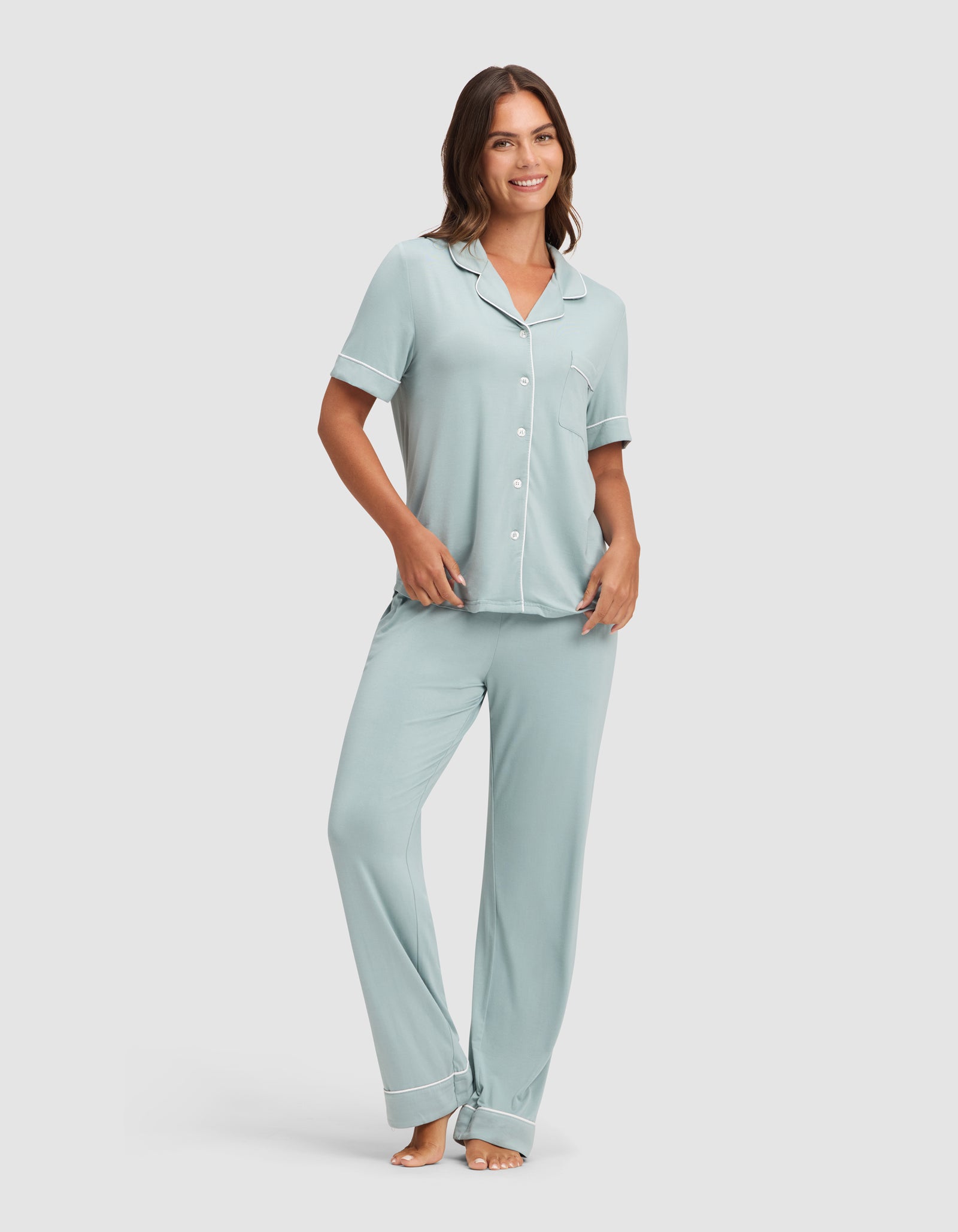A woman smiles barefoot against a plain backdrop, wearing HIDE's Women's Bamboo Stretch-Knit Pajama Pant with a matching light blue, white-trimmed short-sleeve button-up shirt. 