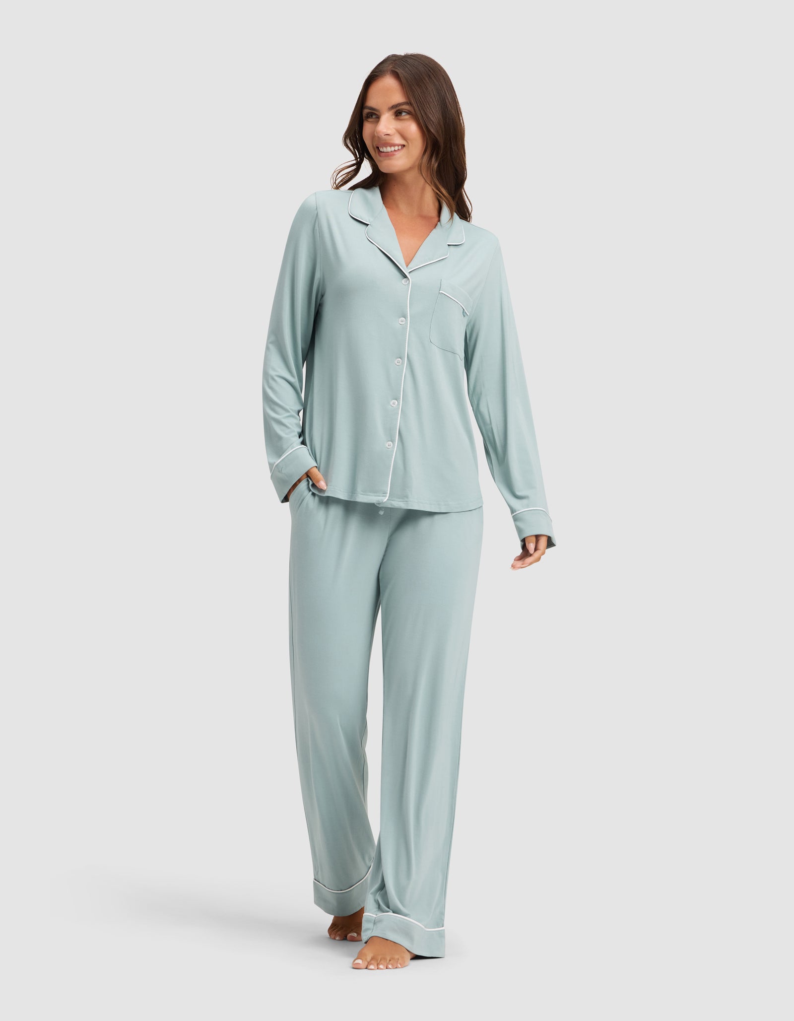A woman smiles while wearing HIDE's Women's Bamboo Stretch-Knit Pajama Pant in light blue with a matching long-sleeve button-up top, barefoot against a plain light background. 