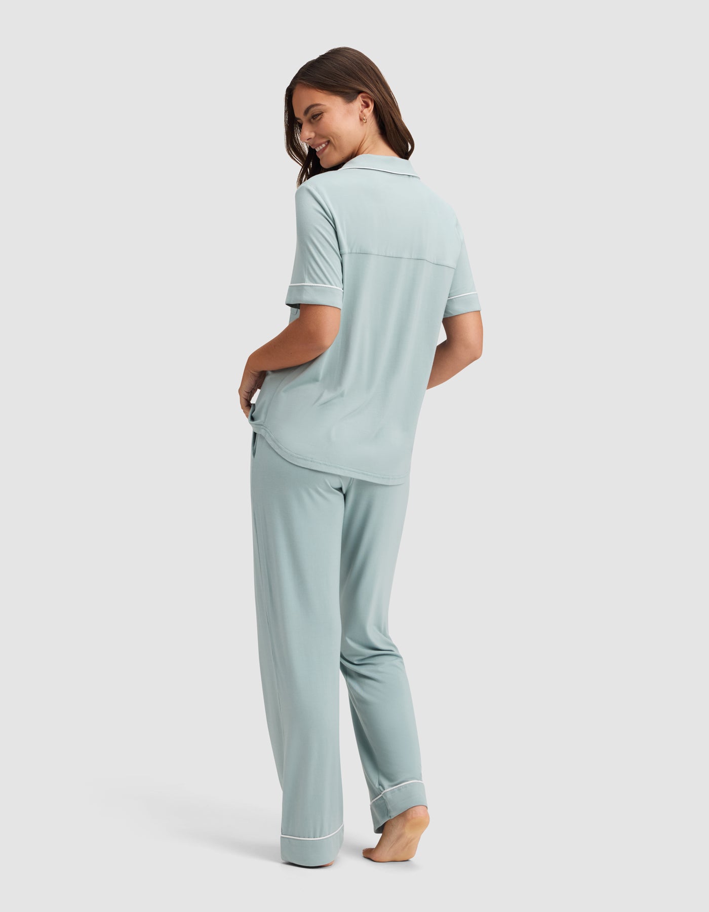 A woman, barefoot and smiling over her shoulder, models the Cozy Earth Women's Bamboo Stretch-Knit Short Sleeve & Pant Pajama Set in light blue with white trim against a simple background. |Color:Brook