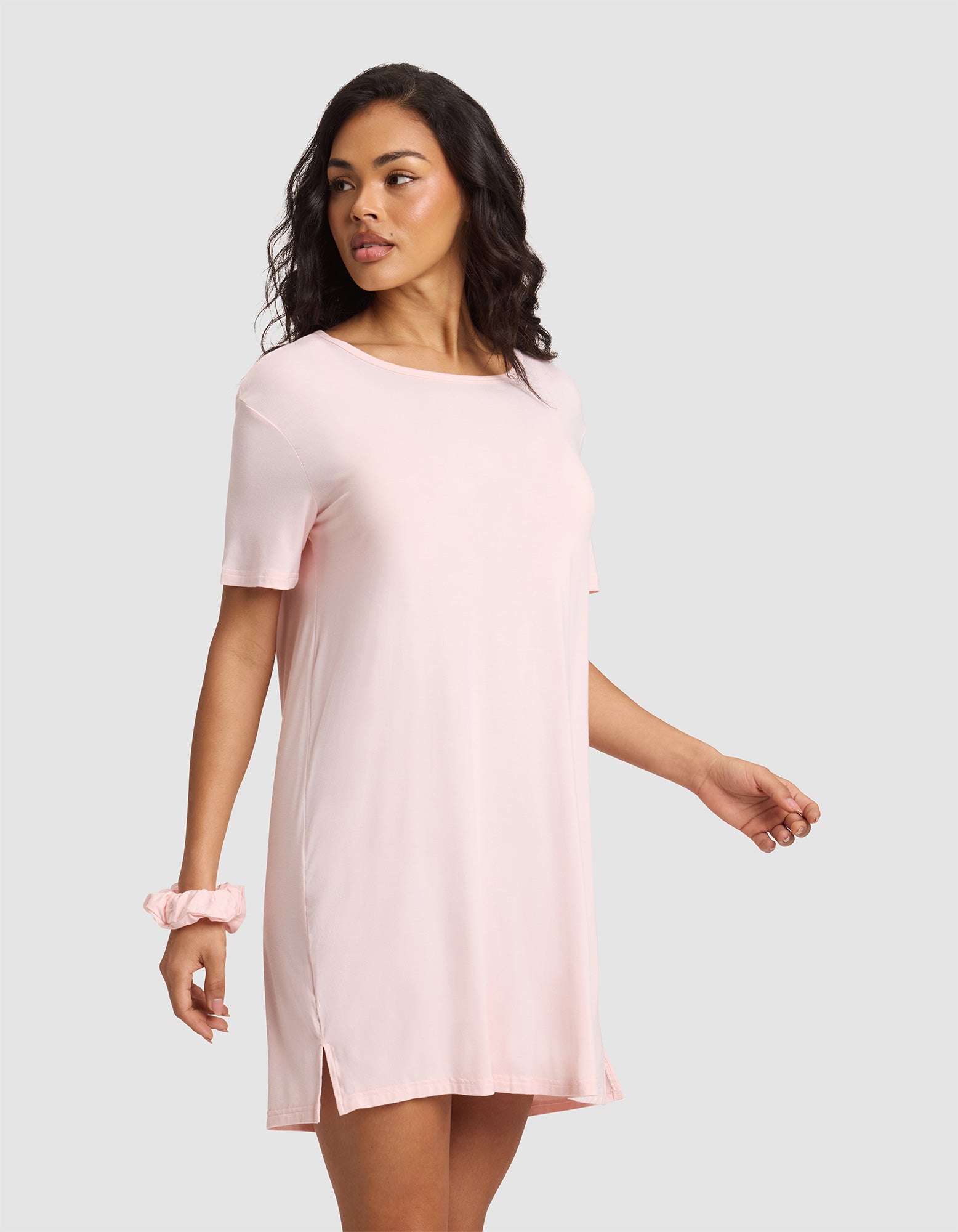 A woman with wavy dark hair and a relaxed expression wears the Cozy Earth Women's Bamboo Stretch-Knit Sleep Dress in light pink. She stands against a plain gray background, with a matching pink scrunchie on her left wrist. 
