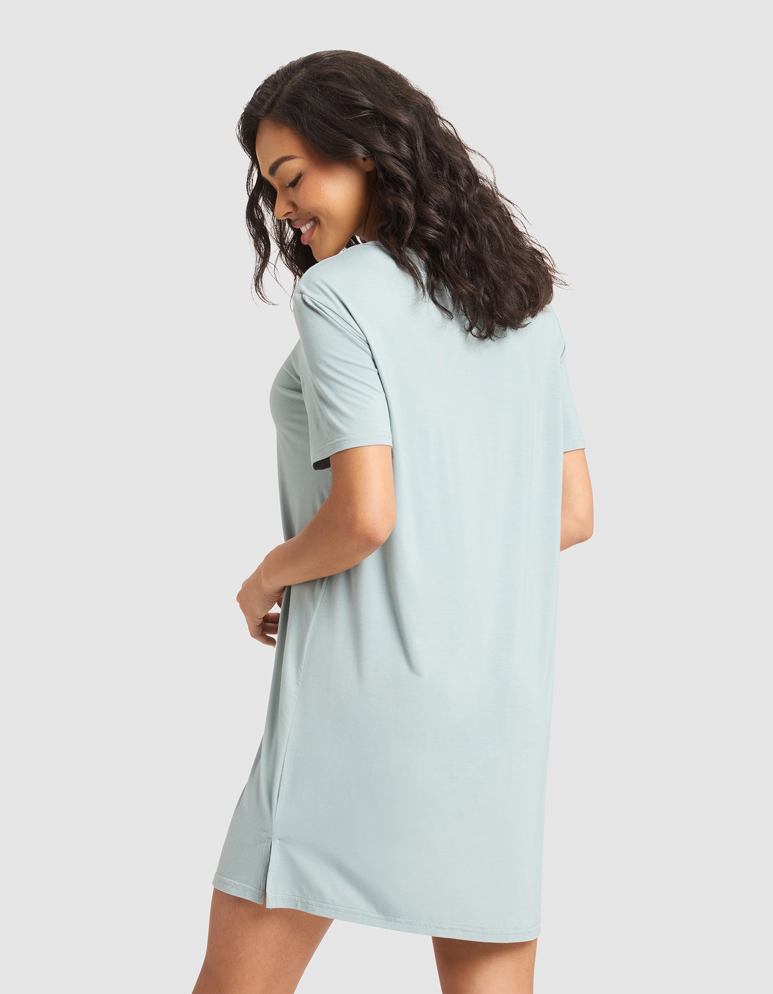 A woman with wavy dark hair smiles and looks down while wearing the light blue Cozy Earth Women's Bamboo Stretch-Knit Sleep Dress. She stands slightly turned away from the camera against a plain background. 