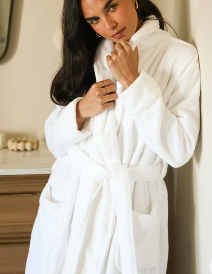 A person with long dark hair stands indoors, wearing a white bathrobe. They are leaning against a wall with one hand adjusting the collar. The background includes a wooden dresser with decorative objects.