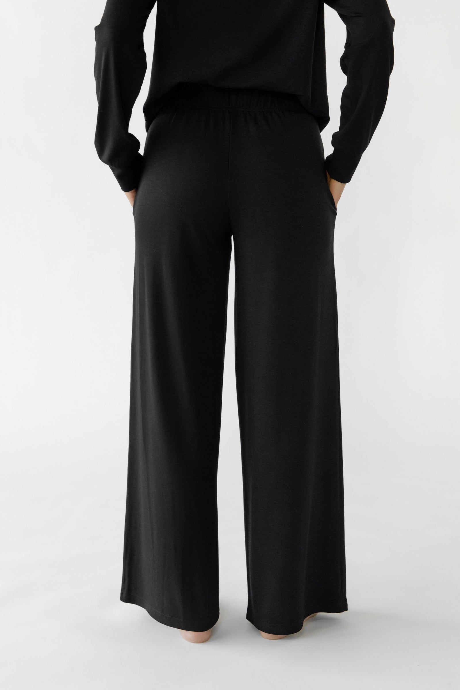 A person stands, facing away while wearing the Women's Ultra-Soft Bamboo Wide Leg Pull On Pant TALL in black from Cozy Earth. The background is a plain, light color. 