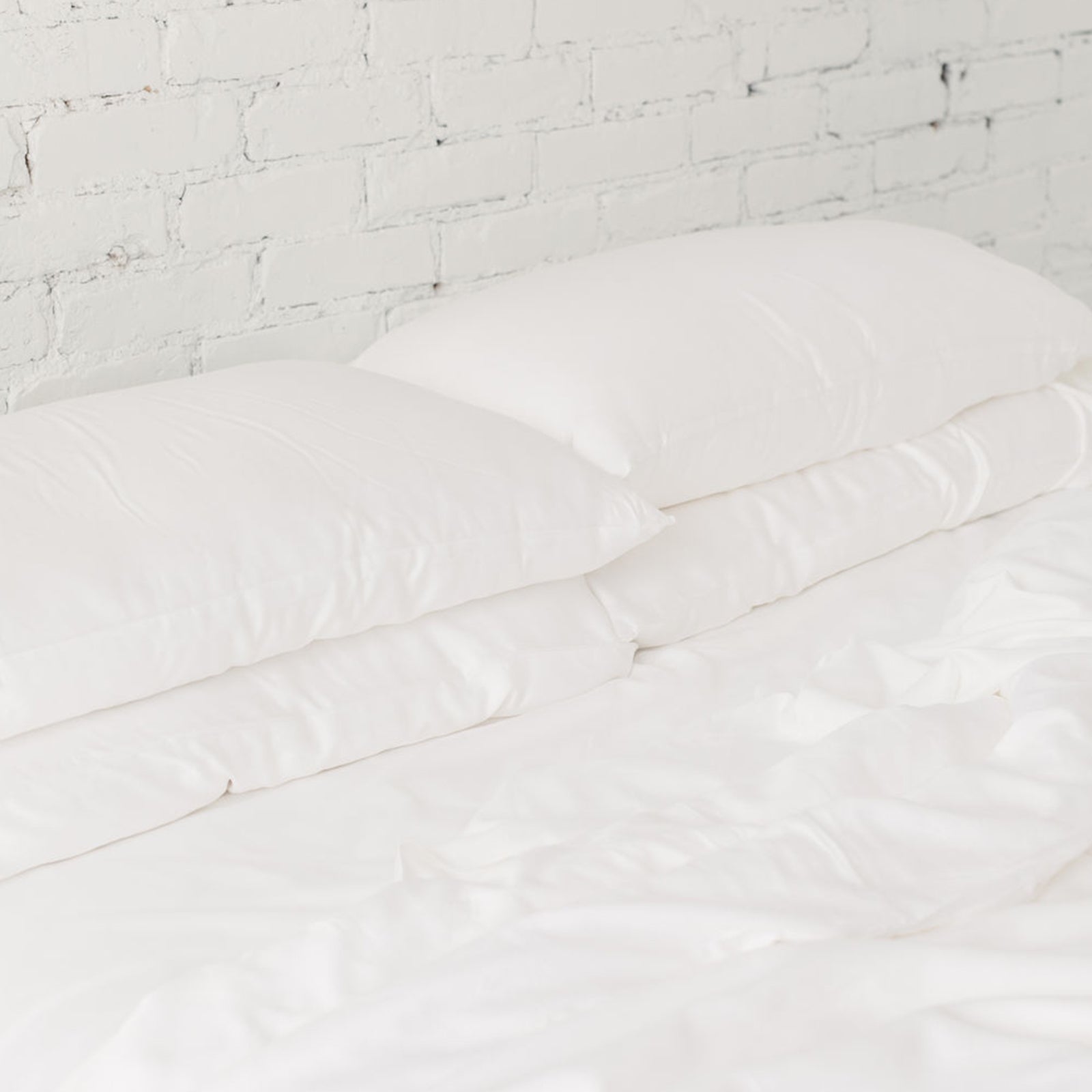 A neatly made bed, featuring Cozy Earth's Silk Pillows and a matching white duvet, is positioned against a white brick wall. The clean and minimalist aesthetic is enhanced by the fluffy and inviting bedding.